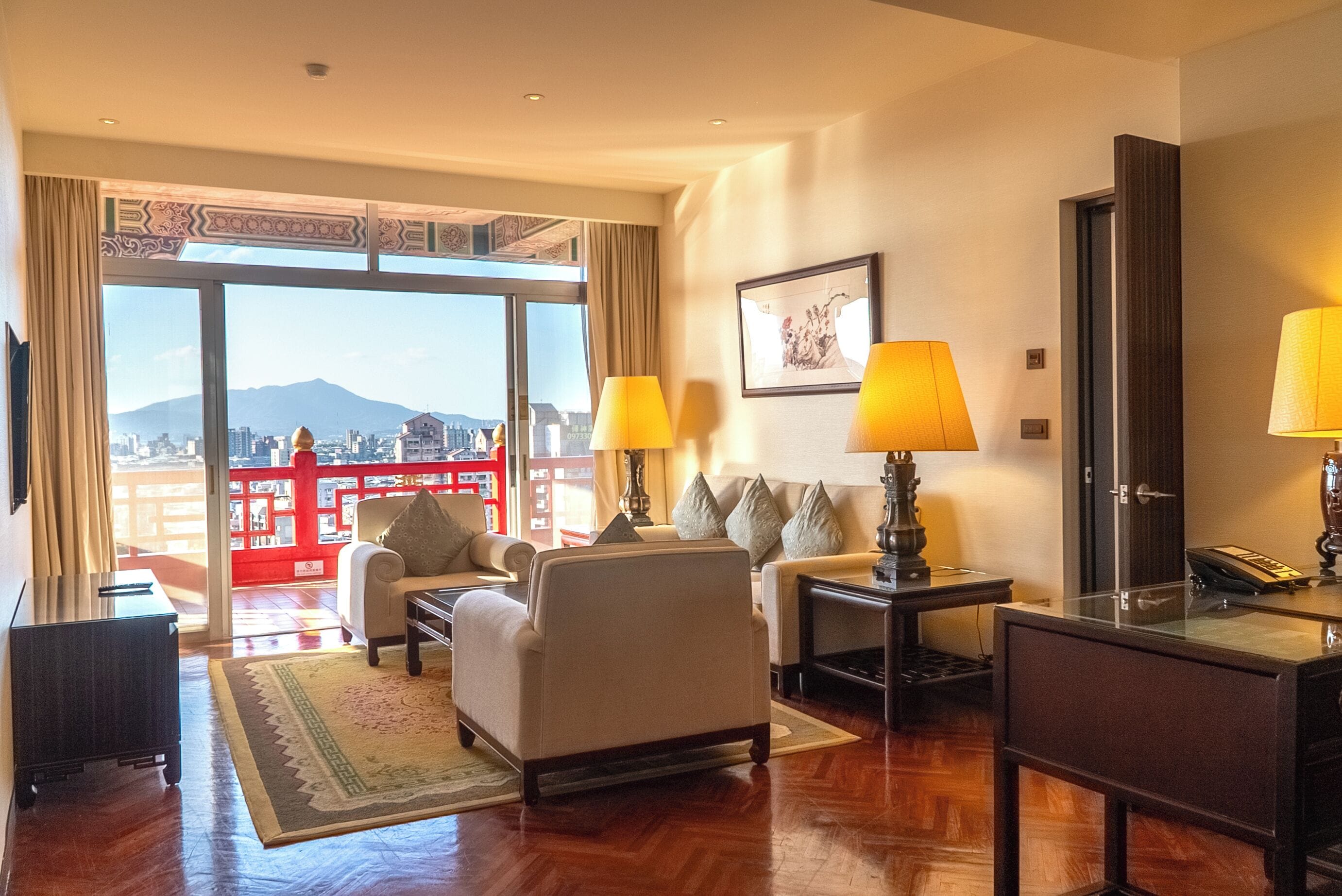 A hotel room with a large window, two lamps, a sofa, a coffee table, and a desk with a telephone.