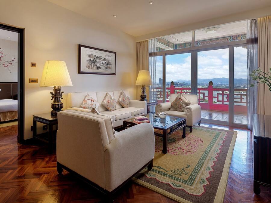A hotel room with a sofa, a bed, a coffee table, and a view of a balcony with a railing.