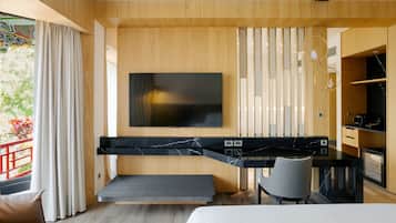 A modern hotel room with a bed, a desk with a chair, a TV mounted on the wall, and a view of greenery outside.