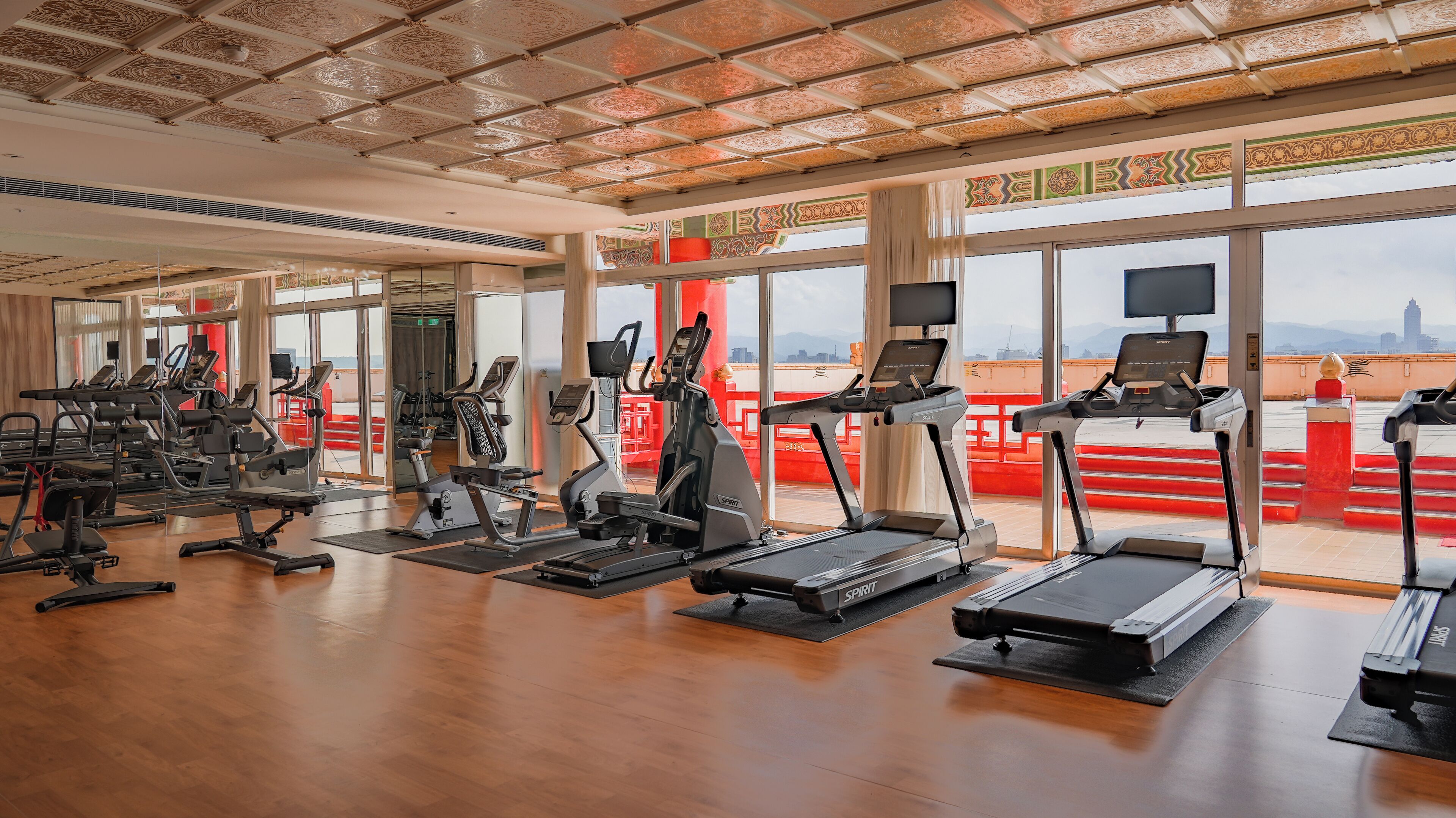 Et rummeligt fitnesscenter med diverse træningsmaskiner, store vinduer og udsigt over bybilledet.