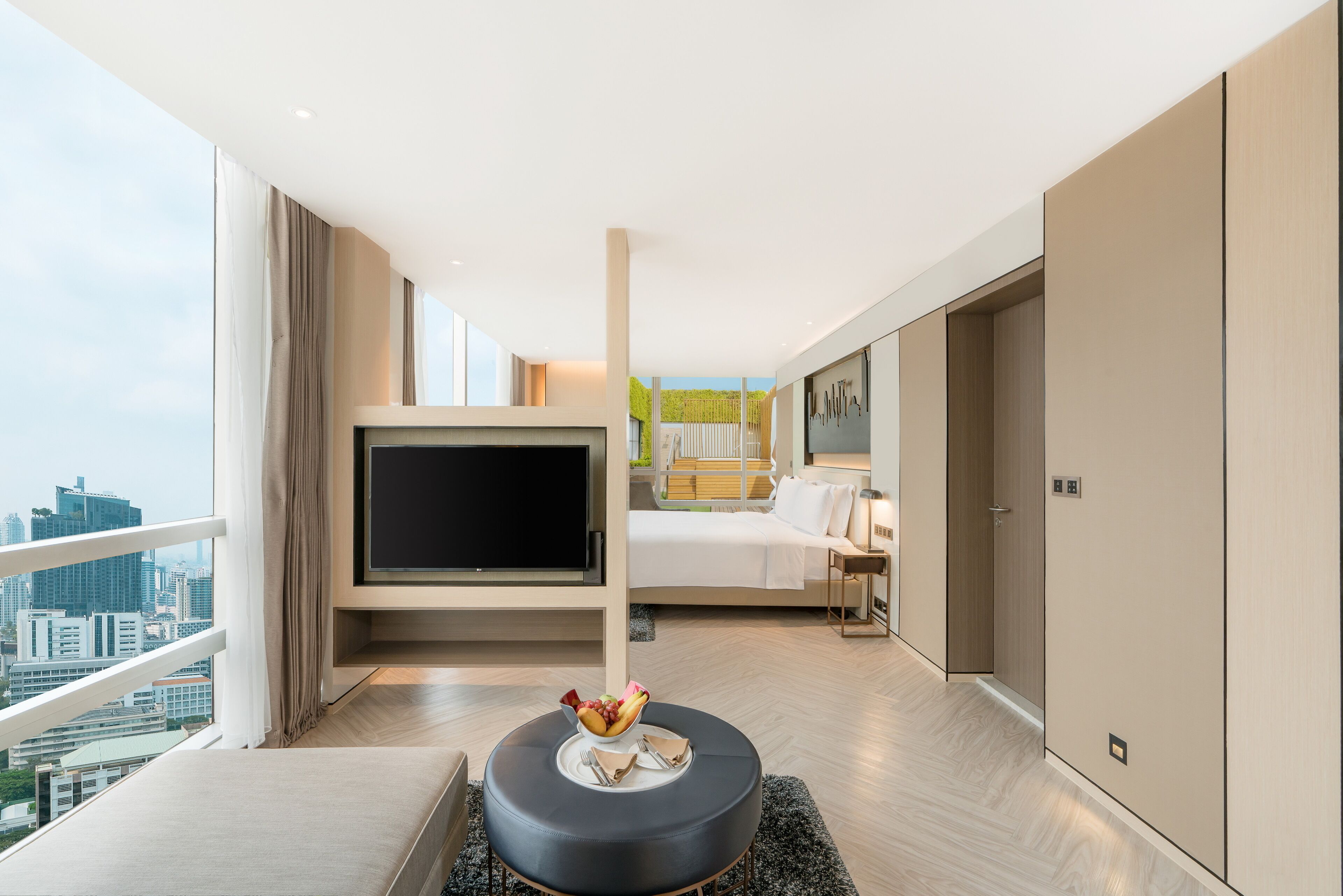 A modern hotel room with a large TV, a bed, a dining area with a table and chairs, and a city view.