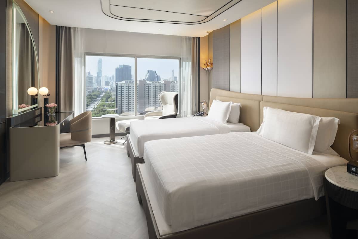 A hotel room with two beds, a sitting area with a chair, a small table, and a large window offering a city view.