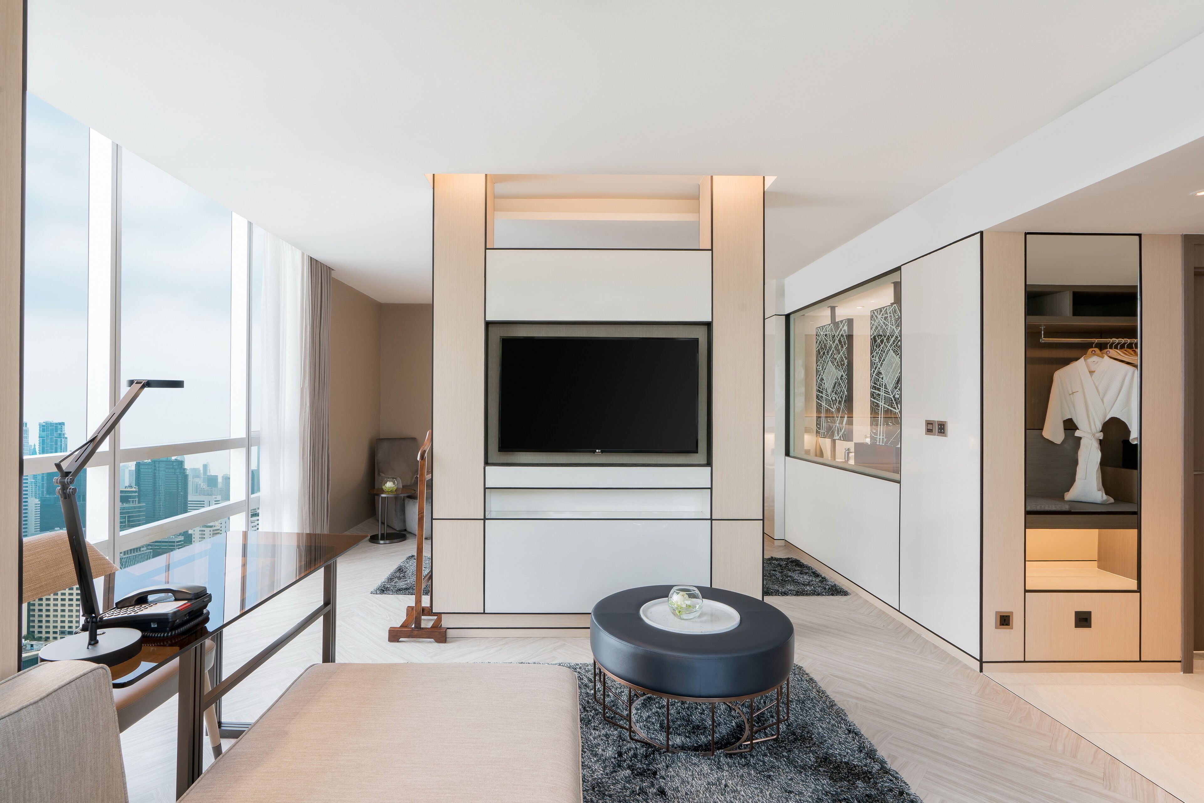 A modern hotel room with a large TV, a seating area with a coffee table, and a view of the city through large windows.