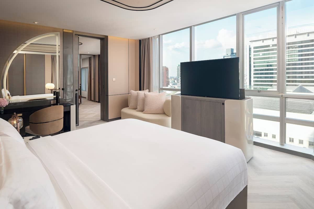 A modern hotel room with a large bed, a desk with a lamp, a television, and a view of the city through large windows.