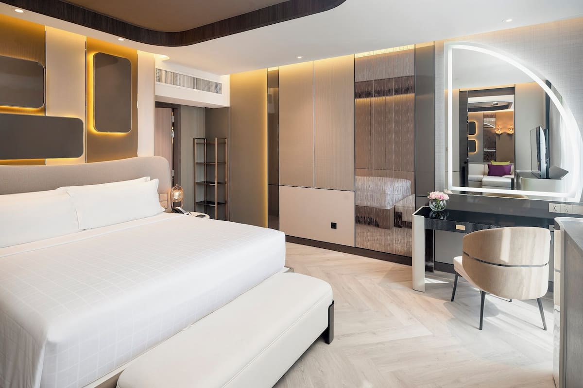 A modern hotel room with a large bed, a vanity with a mirror, and a desk with a chair.