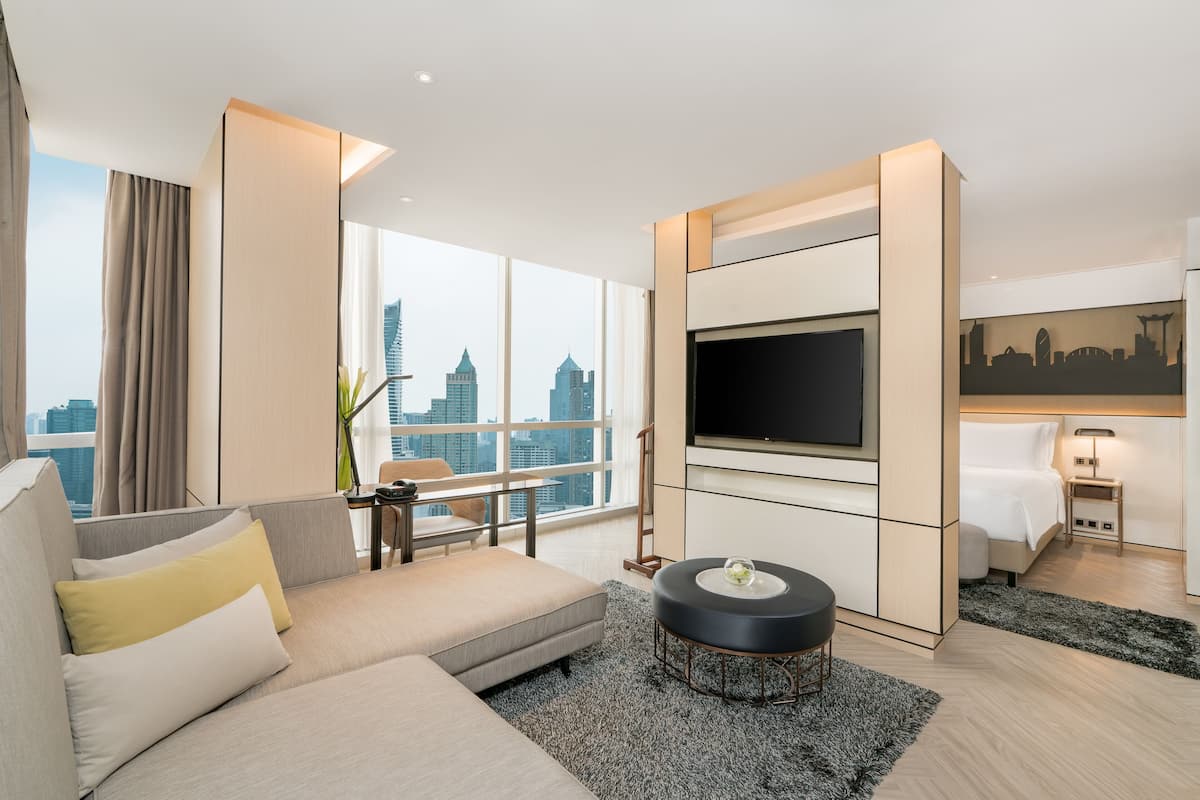 A modern hotel room with a large window, a flat-screen TV, a sofa, a coffee table, and a bed.