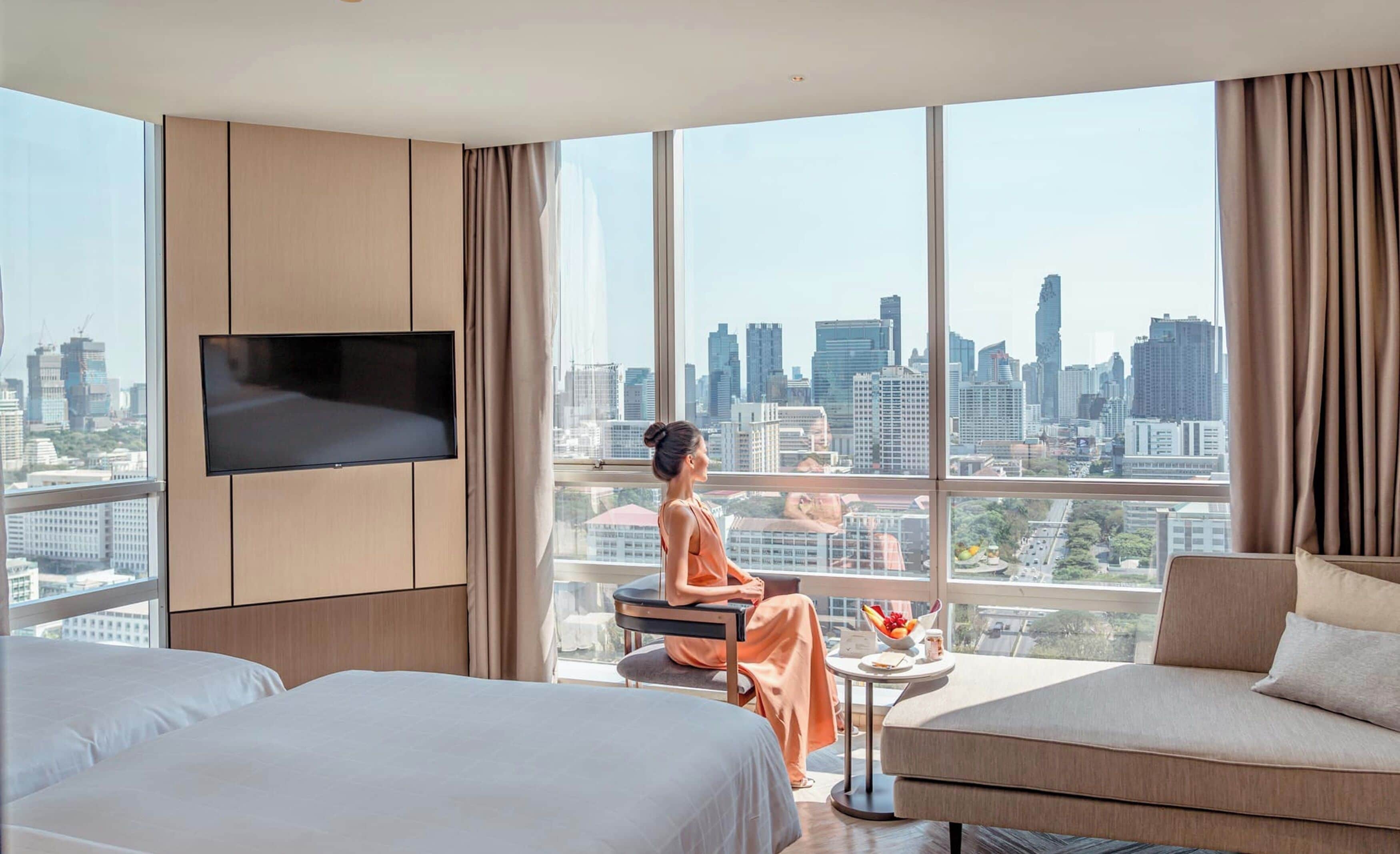 A hotel room with a large window offering a city view, a flat-screen TV mounted on the wall, and a seating area with a table and a bowl of fruit.
