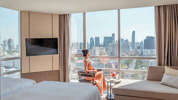 Habitación de hotel con un ventanal grande que ofrece vistas a la ciudad, un televisor de pantalla plana en la pared y un área de descanso con una mesa y un bol de fruta.