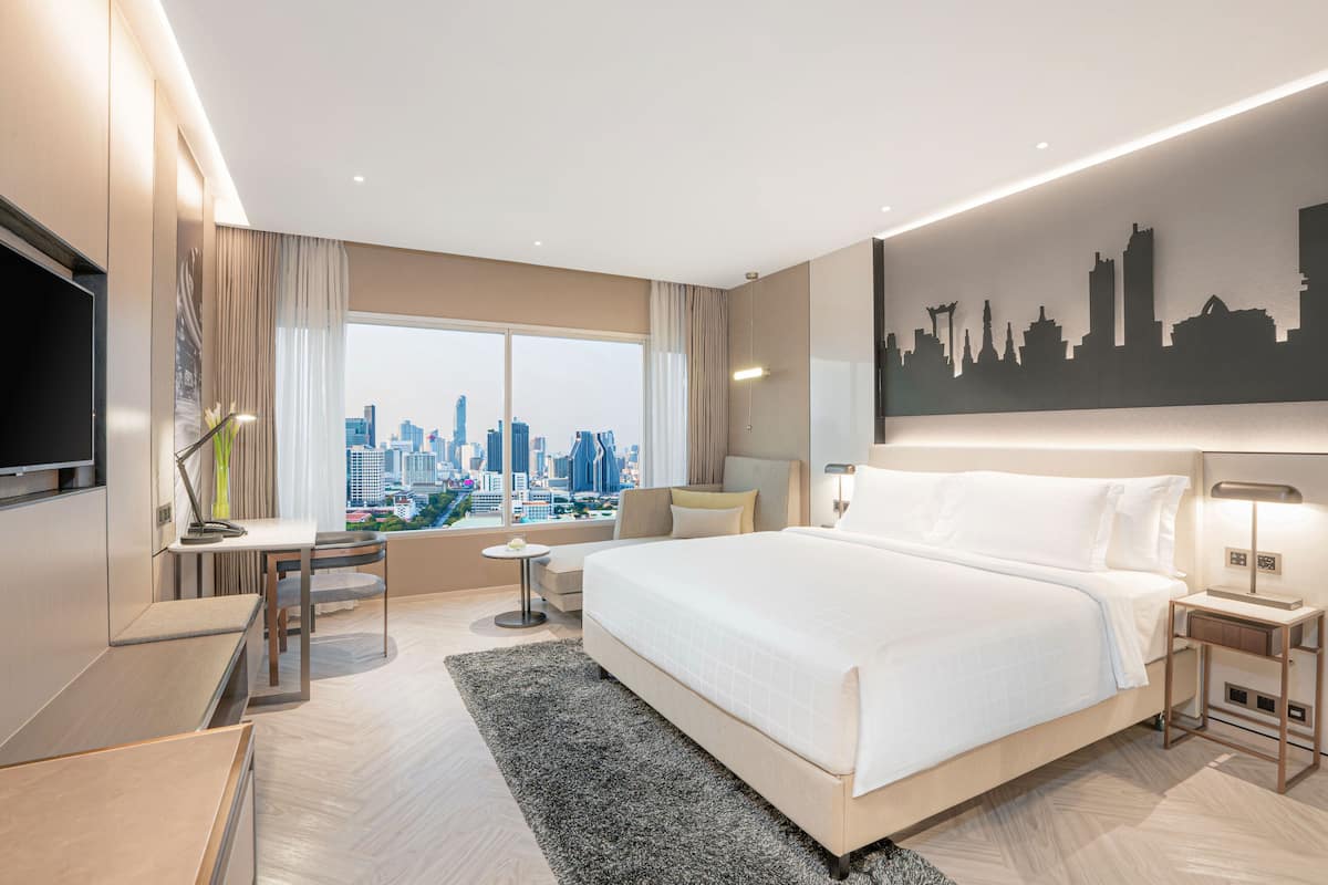 A modern hotel room with a large bed, a desk, a chair, a TV, a city view, and a city skyline artwork on the wall.