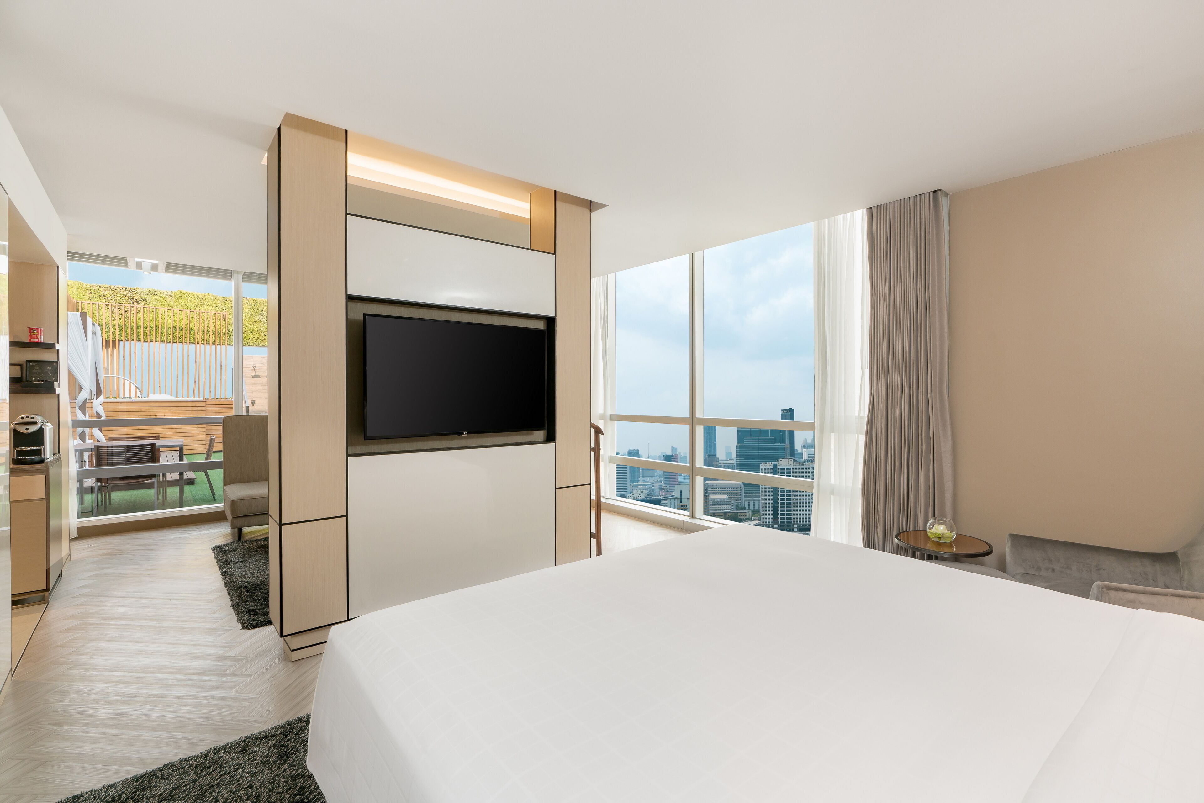 A modern hotel room with a large bed, a flat-screen TV, a balcony with a city view, and a kitchenette.