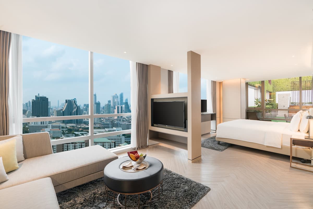 A modern hotel room with a large window, a flat-screen TV, a sofa, a bed, and a small table with fruit.