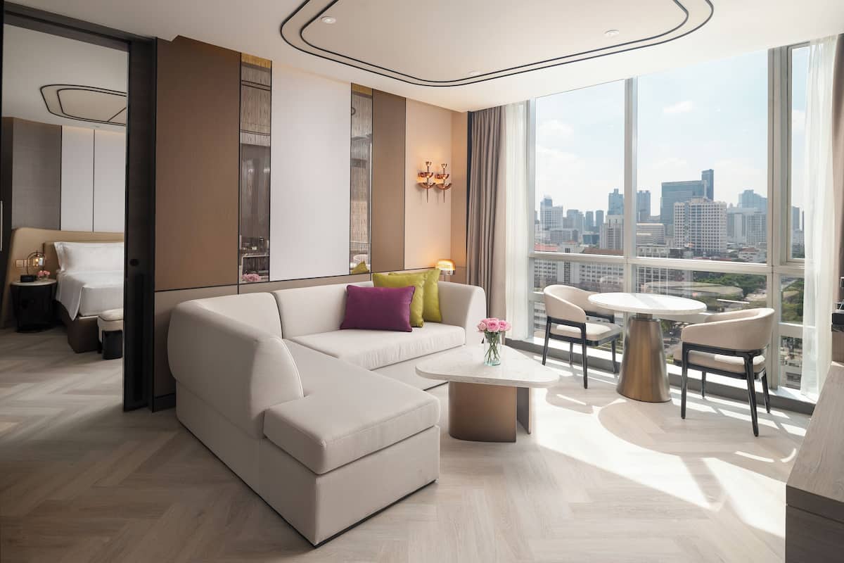 A modern hotel room with a large sofa, a dining area, and a city view.