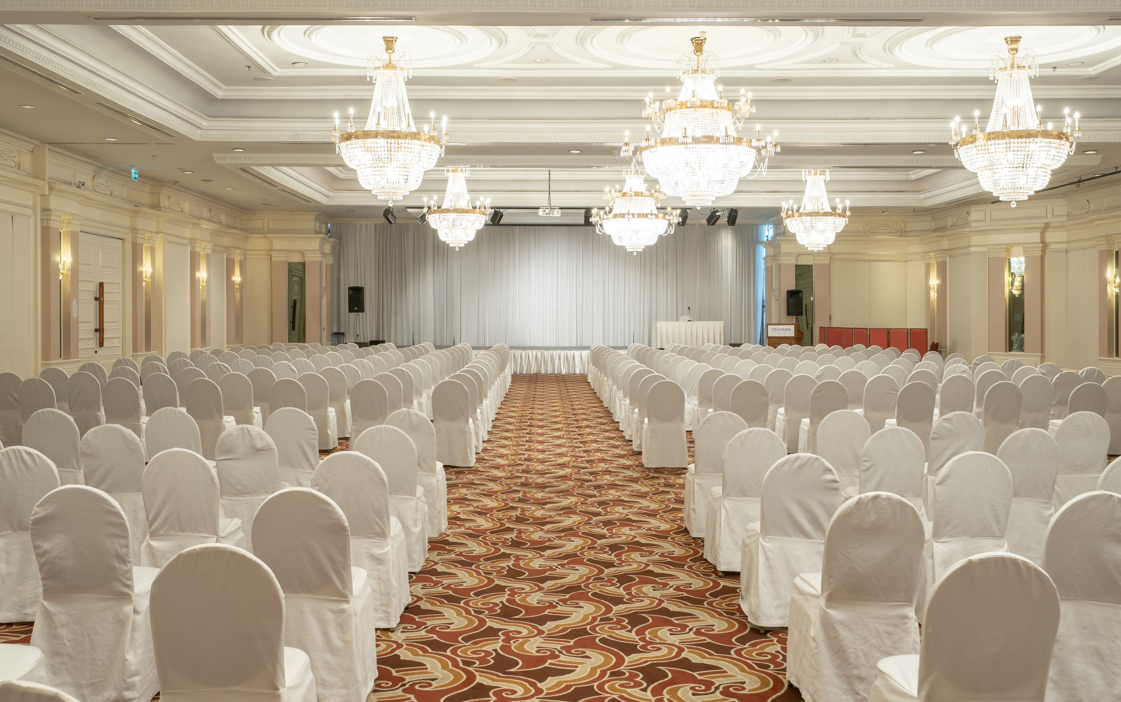 Un amplio salón de conferencias con filas de sillas cubiertas de blanco, una alfombra estampada y varios candelabros.