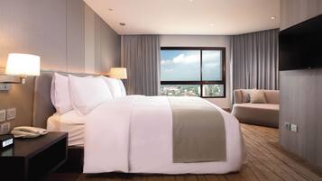 Habitación de hotel con una cama grande, un sofá, un televisor, una ventana con vistas a la ciudad y un escritorio con teléfono.