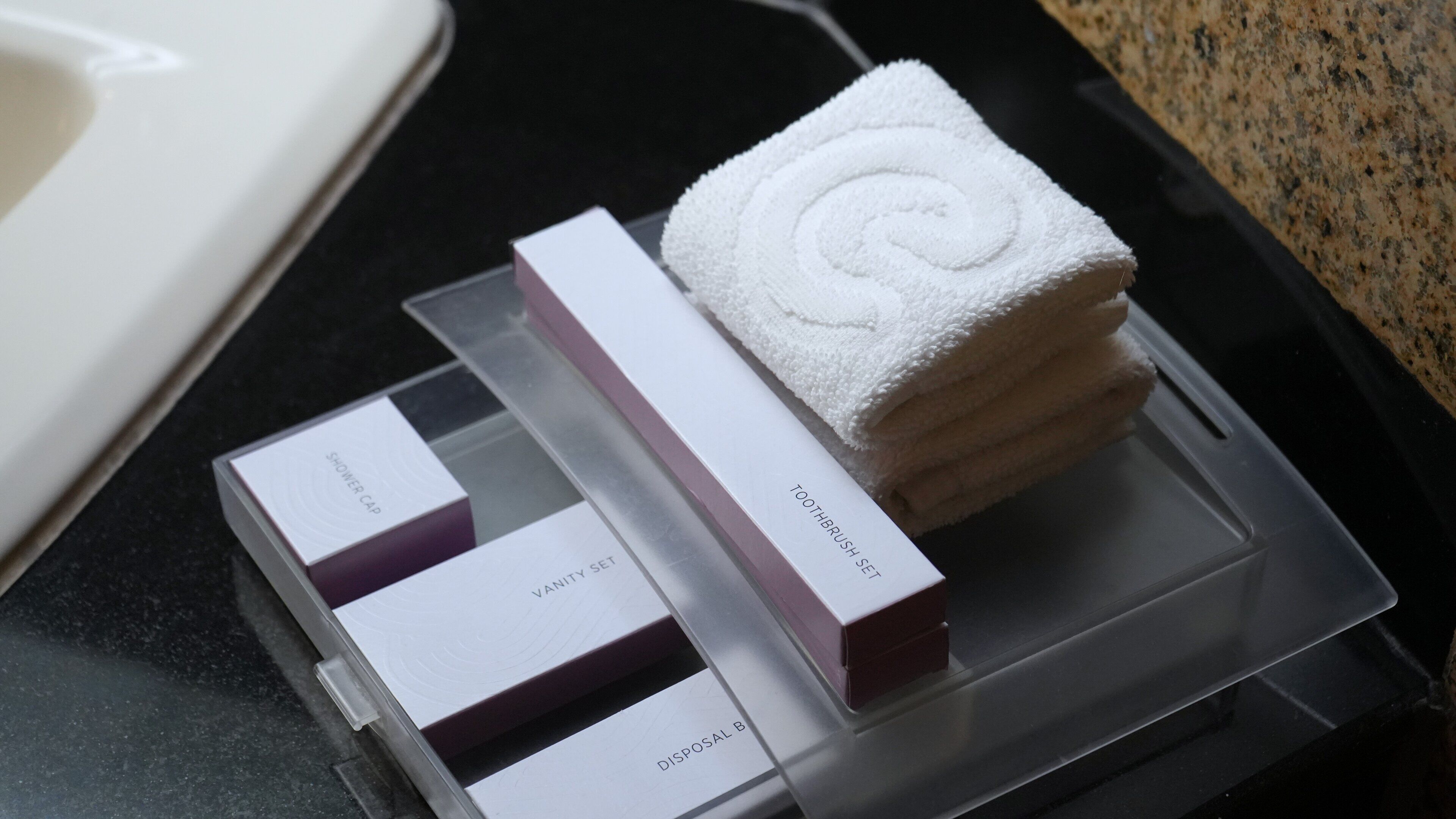 A clear plastic tray with a white towel, a toothbrush set, a vanity set, and a disposal box.