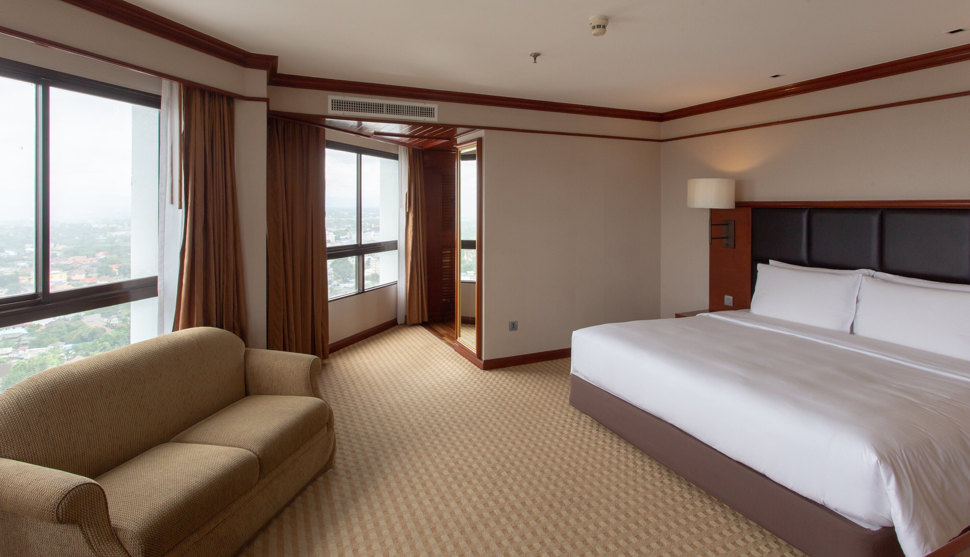 A hotel room with a large bed, a sofa, a window with curtains, and an air conditioning unit.