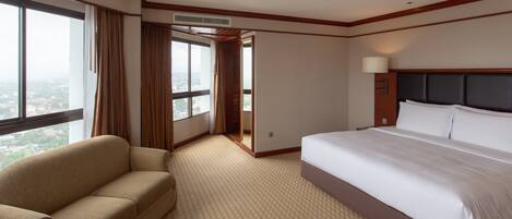 A hotel room with a large bed, a sofa, a window with curtains, and an air conditioning unit.