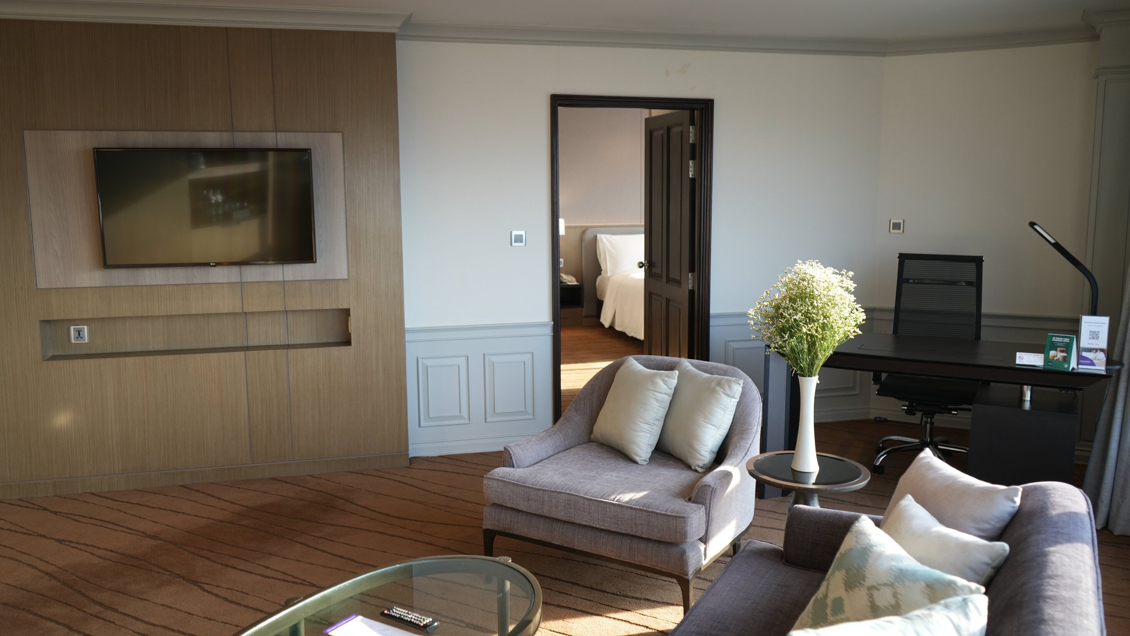 Ein modernes Hotelzimmer mit einem an einer Holzvertäfelung montierten Flachbildfernseher, einem grauen Sofa mit weißen Kissen, einem runden Glastisch und einem kleinen Beistelltisch mit einer Blumenvase.