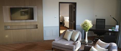 Una habitación de hotel moderna con una televisión de pantalla plana montada en una pared de paneles de madera, un sofá gris con cojines blancos, una mesa de centro redonda de cristal y una mesita auxiliar con un jarrón de flores.
