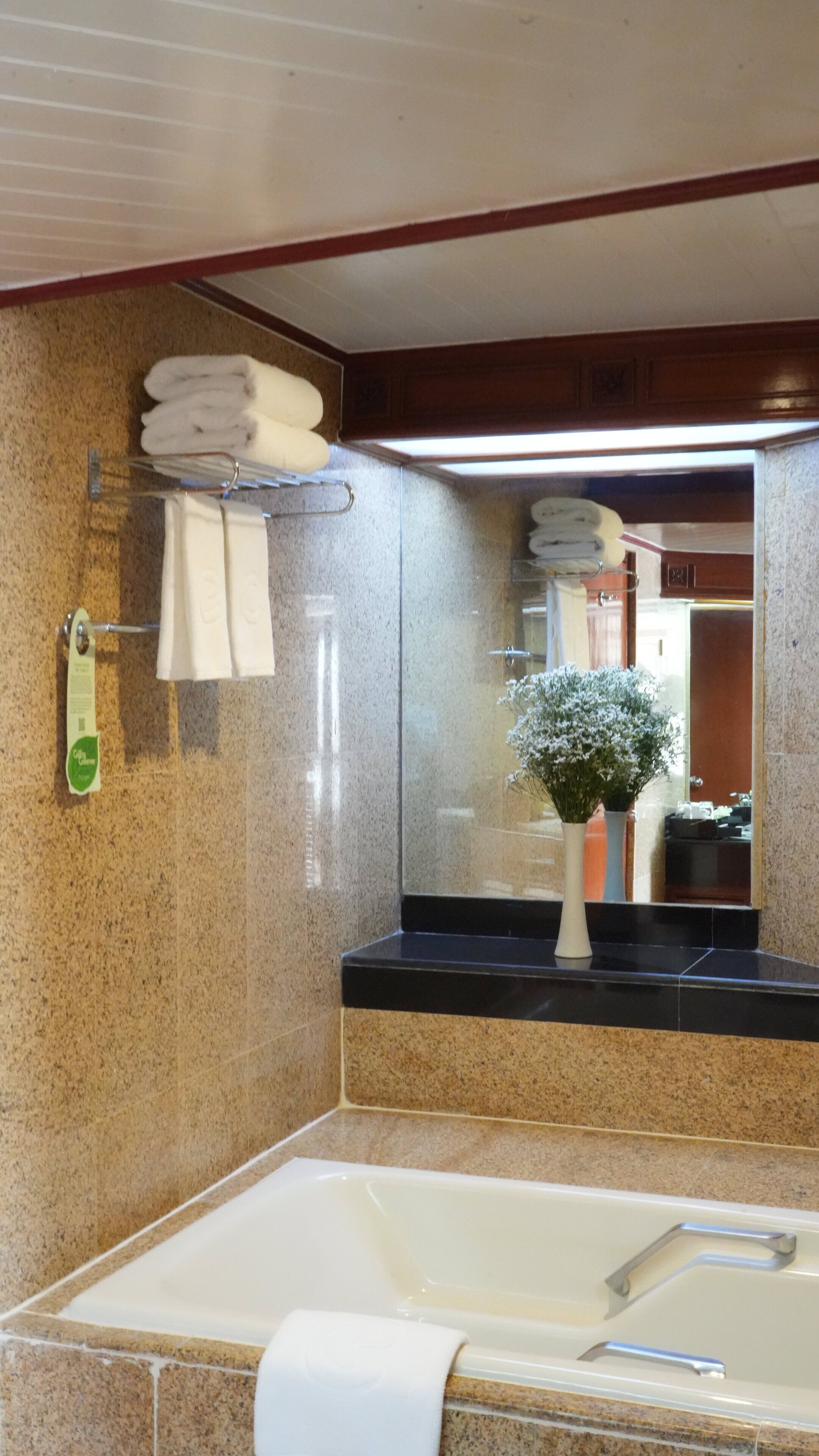 A bathroom with a granite countertop, a white bathtub, a mirror, and a flower vase.
