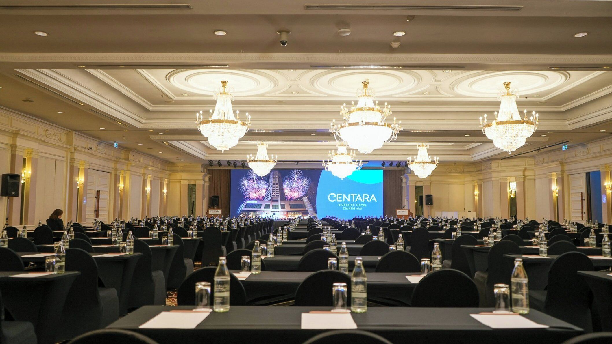 A large conference room with rows of tables, water bottles, and a stage with a screen displaying 'CENTARA RIVERSIDE HOTEL CHIANG MAI'.