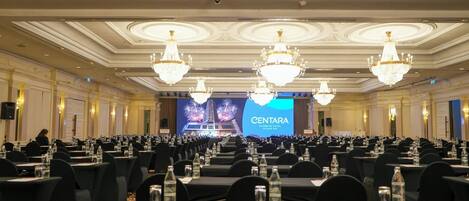 Uma grande sala de conferências com fileiras de mesas, garrafas de água e um palco com uma tela exibindo 'CENTARA RIVERSIDE HOTEL CHIANG MAI'.