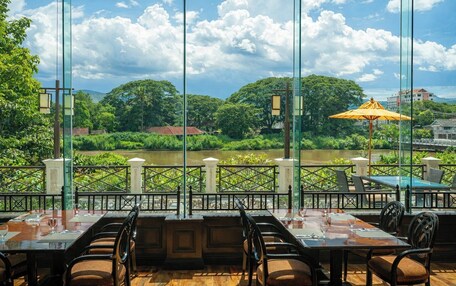 A restaurant with outdoor seating overlooking a river, featuring glass railings and a view of greenery and buildings.