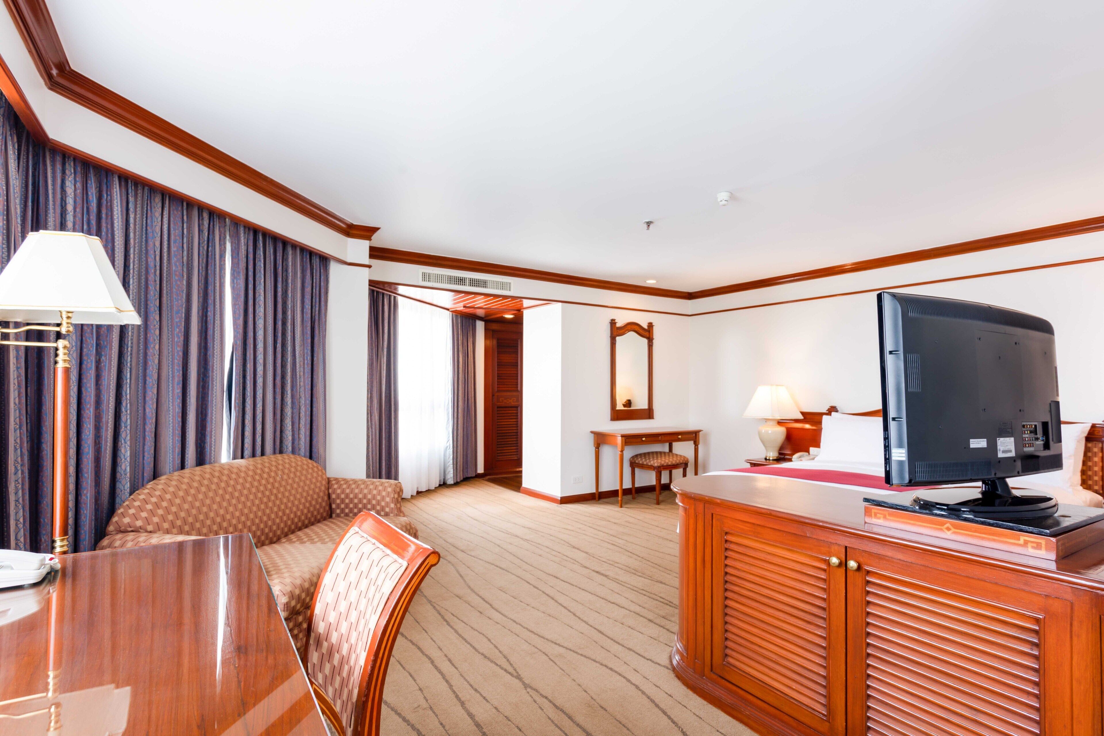 A hotel room with a wooden desk, a television, a sofa, a dining table, and a mirror.