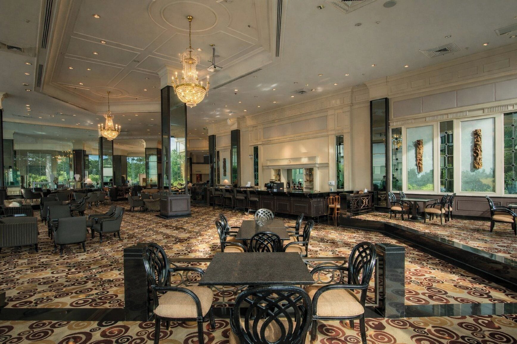 A spacious hotel lobby with a patterned carpet, multiple seating areas, a bar, and large windows offering views.