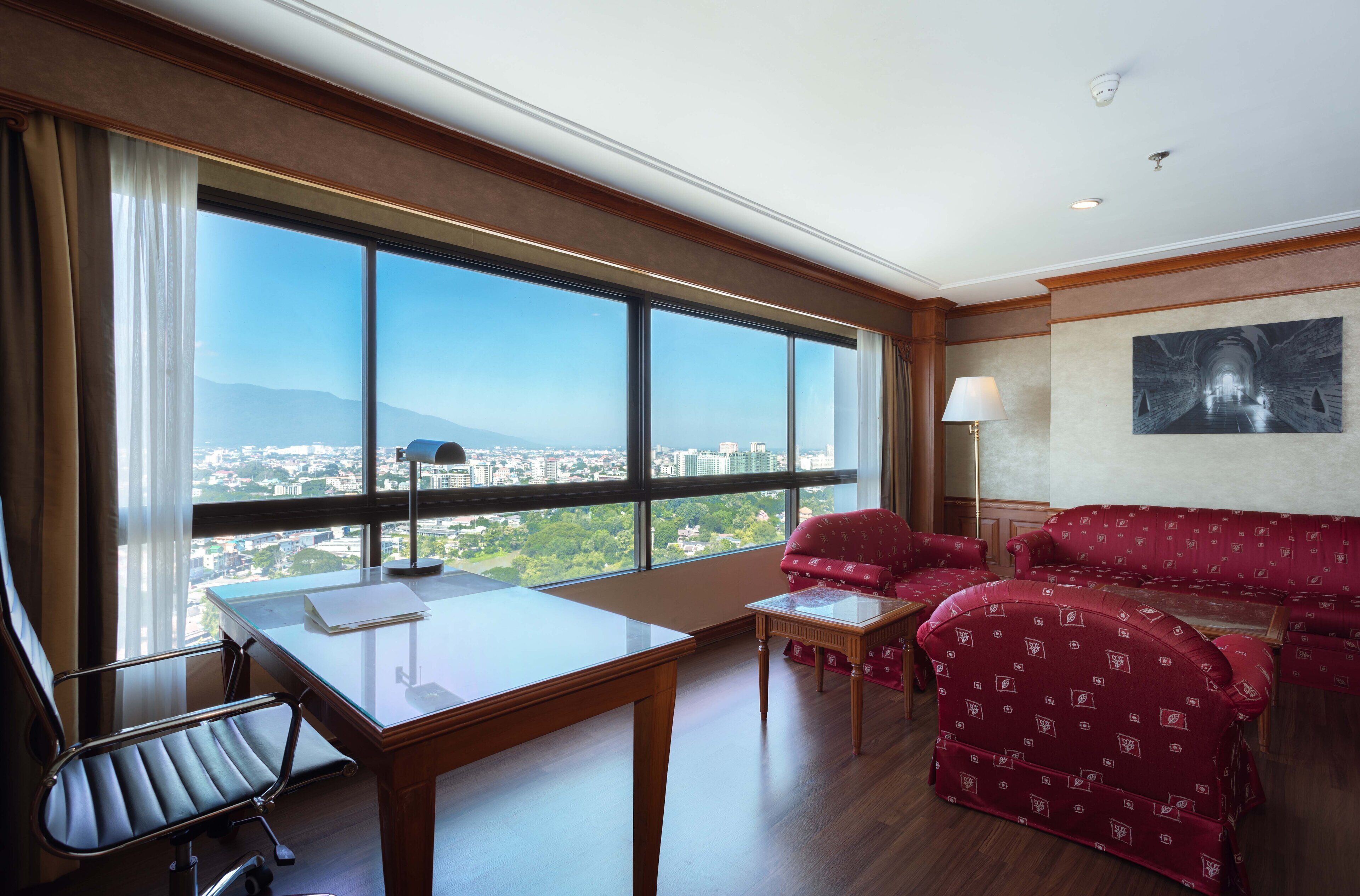 A hotel room with a large window, a desk, a chair, a sofa, and a lamp.