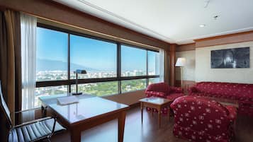 A hotel room with a large window, a desk, a chair, a sofa, and a lamp.