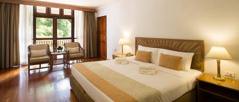 A hotel room with a large bed, two bedside tables with lamps, a wooden headboard, a window with curtains, and a seating area with chairs.