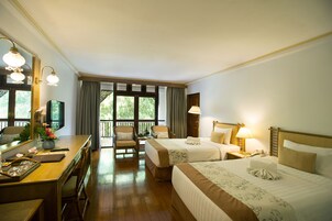 Habitación de hotel con dos camas, un escritorio, una silla, una lámpara y un ventanal grande con cortinas.