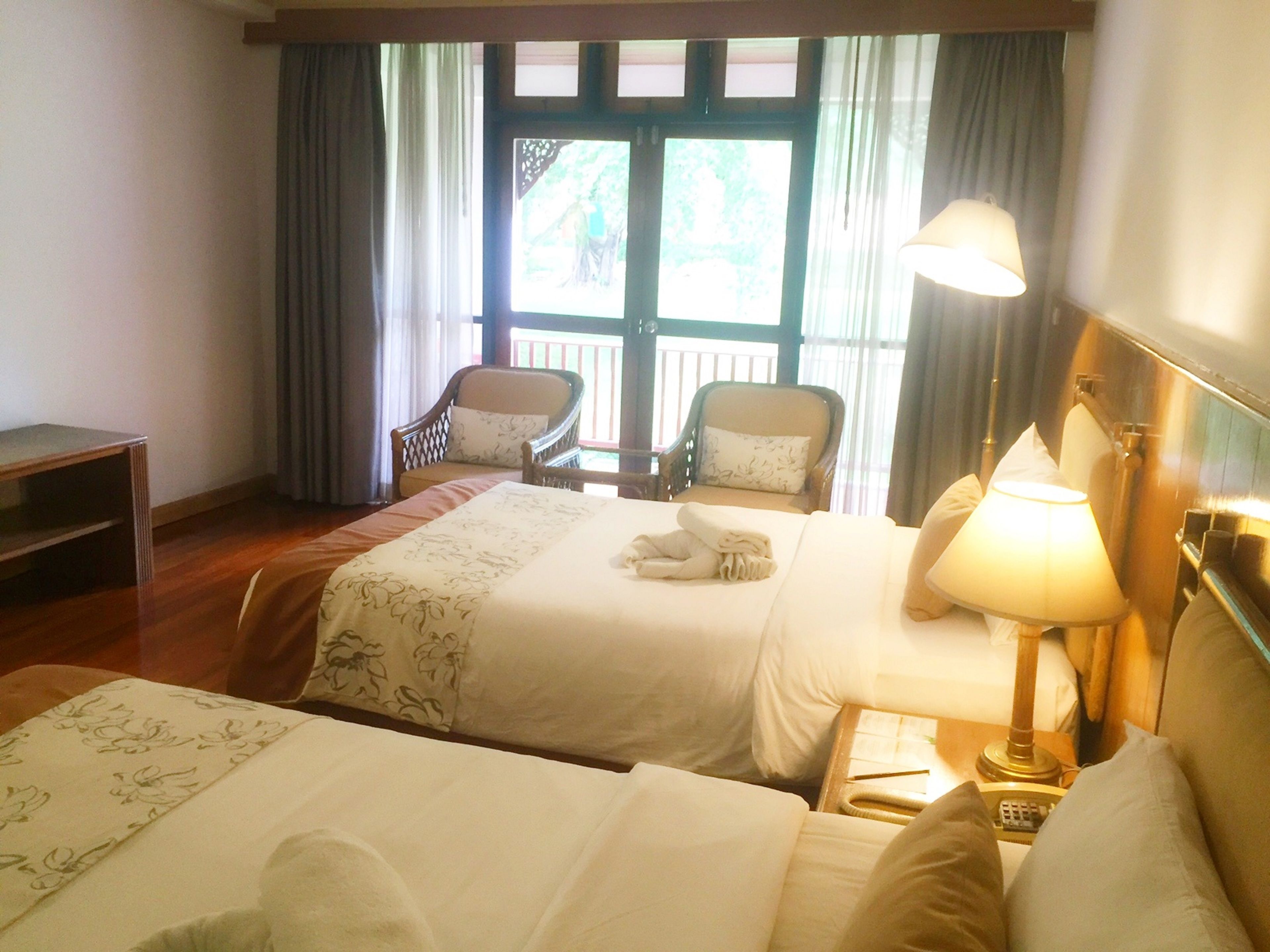 Habitación de hotel con dos camas, un escritorio, dos sillas, una lámpara y vistas al exterior a través de una ventana.