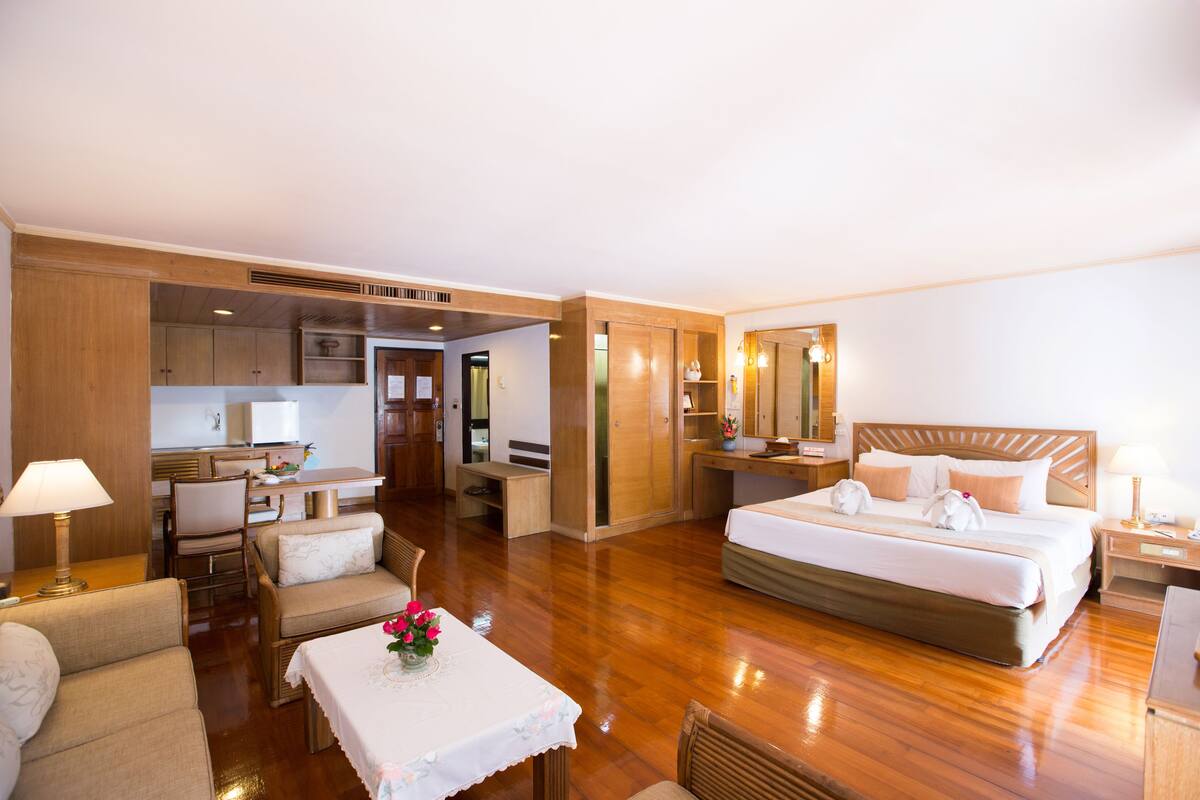 Ein Hotelzimmer mit einem Bett, einem Schreibtisch, einem Sessel, einem Tisch mit Blumenvase und einer Küchenzeile im Hintergrund.
