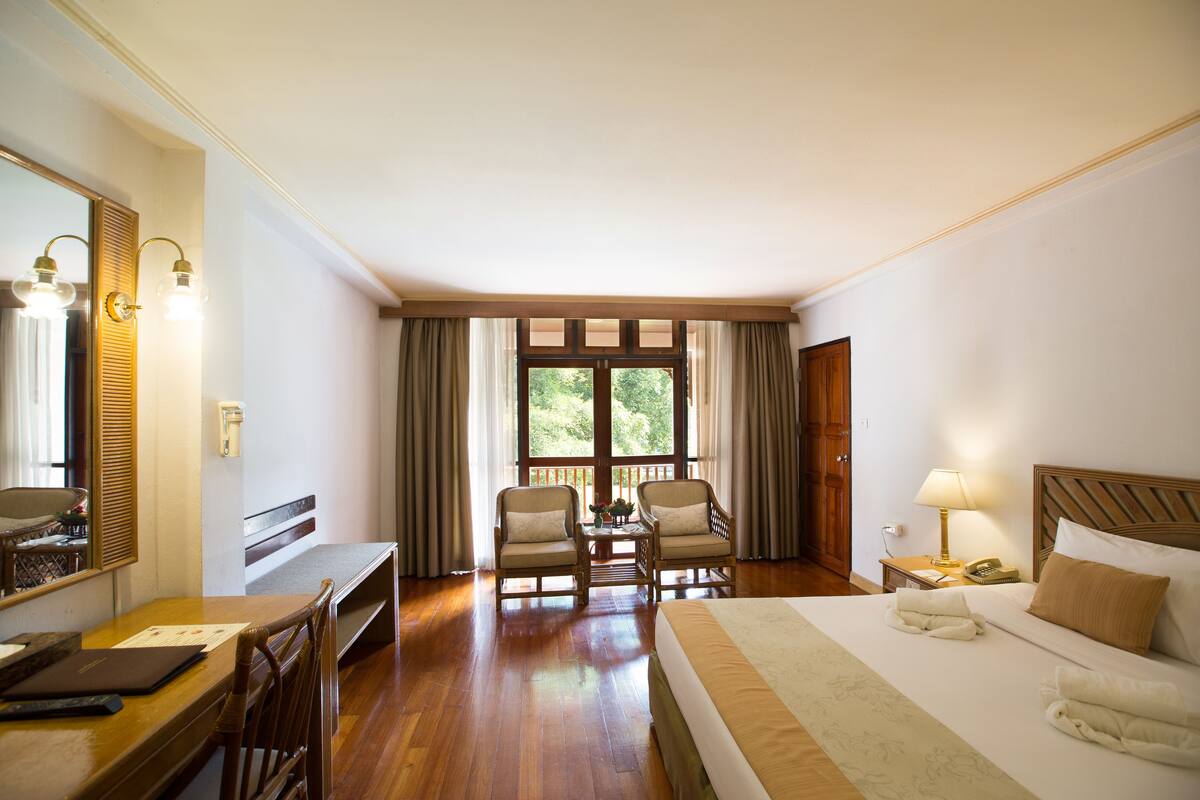 Ein Hotelzimmer mit Bett, Schreibtisch, Stühlen, einem fenster mit Vorhängen und einem Nachttisch mit Lampe.