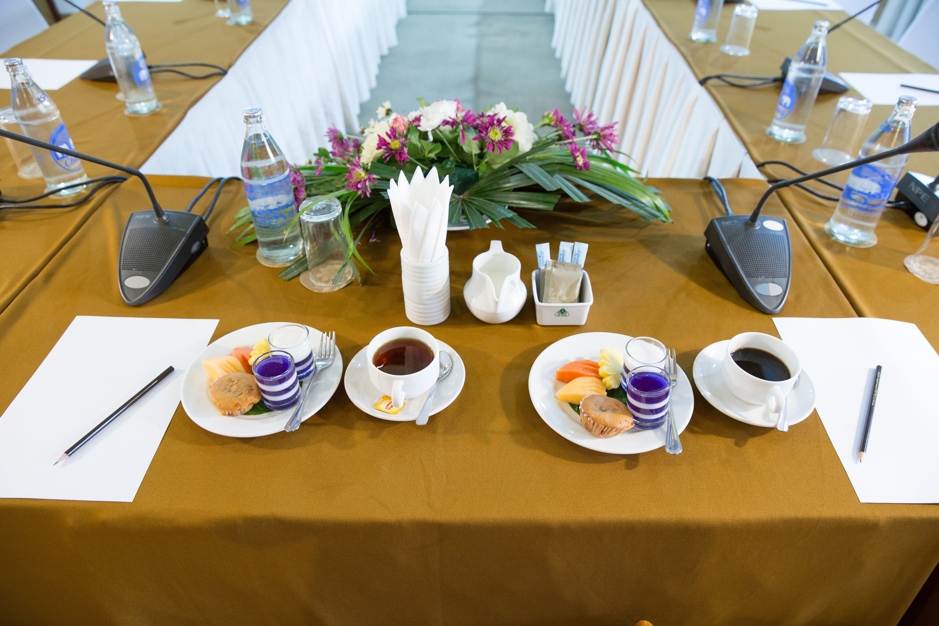 A conference table with microphones, water bottles, a flower arrangement, and plates of food and beverages.