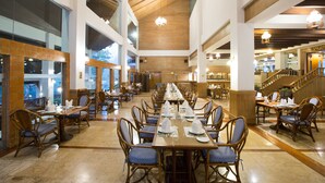 A spacious dining area with high ceilings and natural light from large windows. Tables are set with white napkins and plates, and there are multiple dining tables arranged in rows.