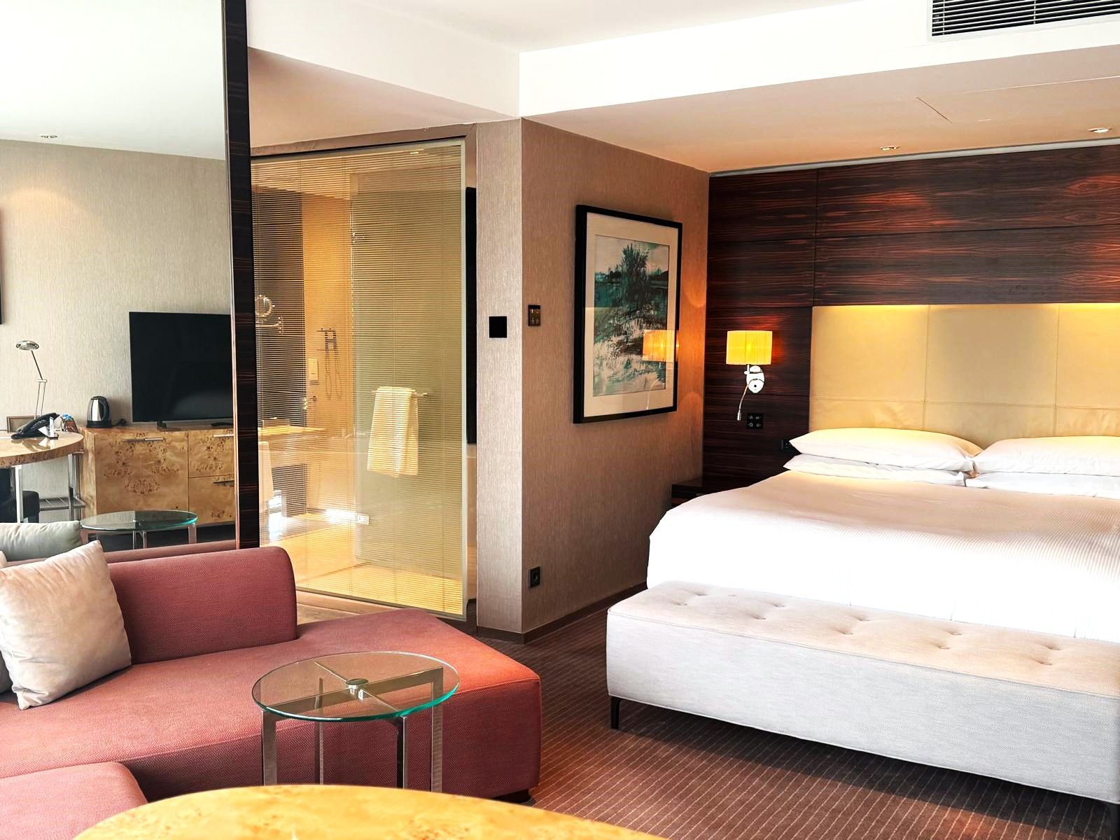 A hotel room with a bed, a sofa, a glass table, a TV, and a painting on the wall.