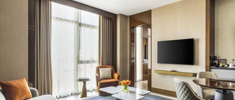 A hotel room with a flat-screen TV, a sofa, a coffee table, a dining area, and a view of the city through large windows.