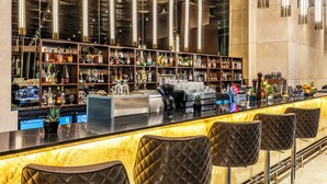 Un bar con mostrador de mármol, varias estanterías llenas de botellas de licor y tres taburetes acolchados.
