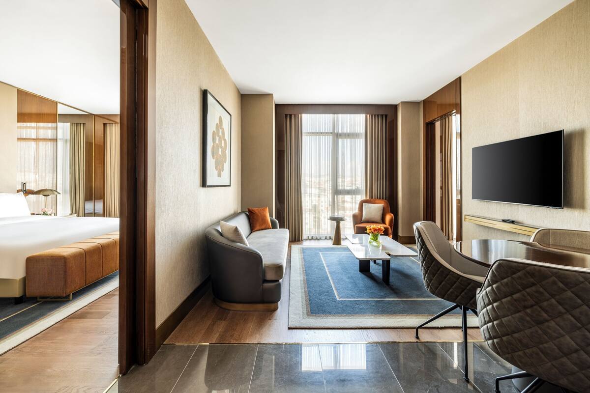 A modern hotel room with a bed, sofa, armchair, and a flat-screen TV.