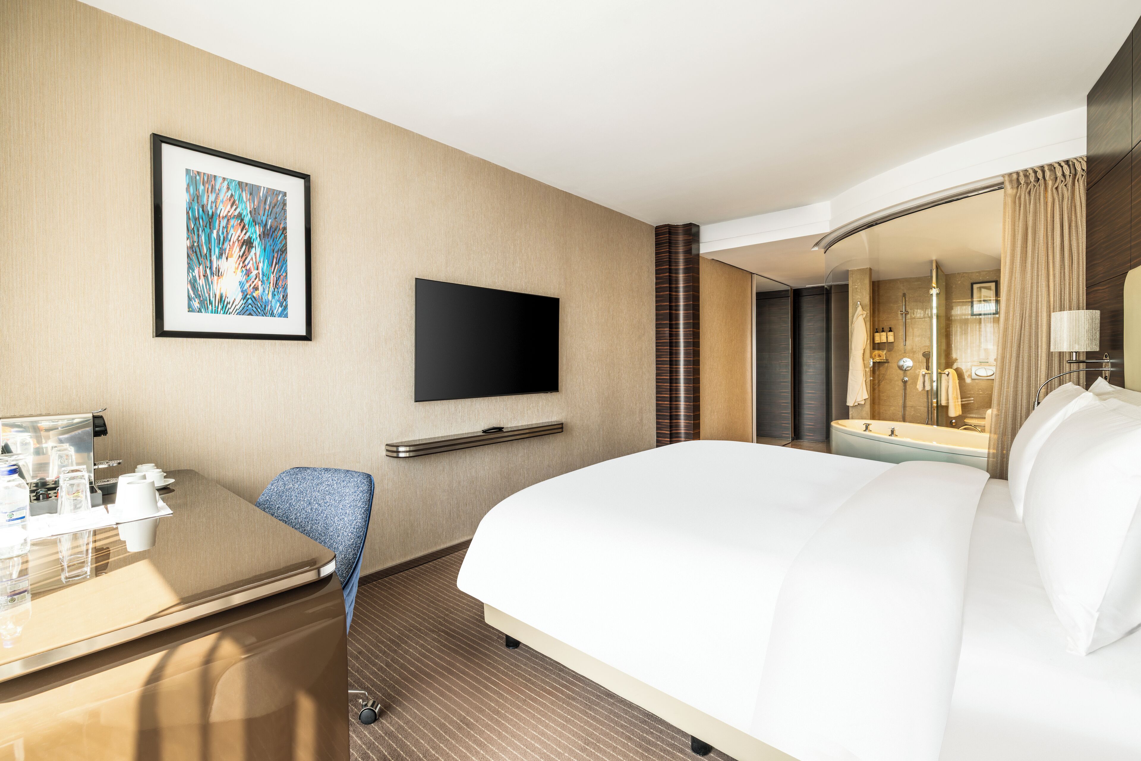 A hotel room with a large bed, a desk with a chair, a flat-screen TV mounted on the wall, and a bathroom visible through a glass partition.
