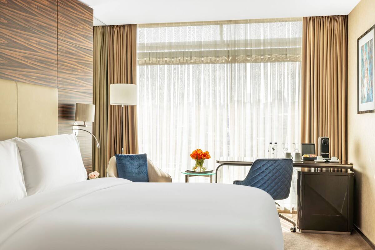 A hotel room with a large bed, a desk with a computer, a chair, and a small table with a vase of flowers.