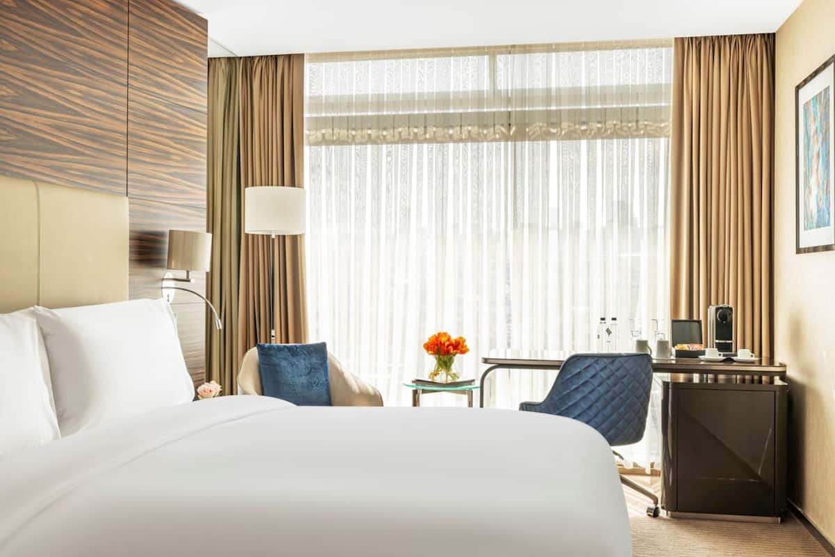 A hotel room with a large bed, a desk with a computer, a chair, and a small table with a vase of flowers.
