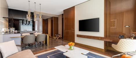 A modern hotel room with a dining area, a flat-screen TV mounted on the wall, and a seating area with a coffee table and chairs.