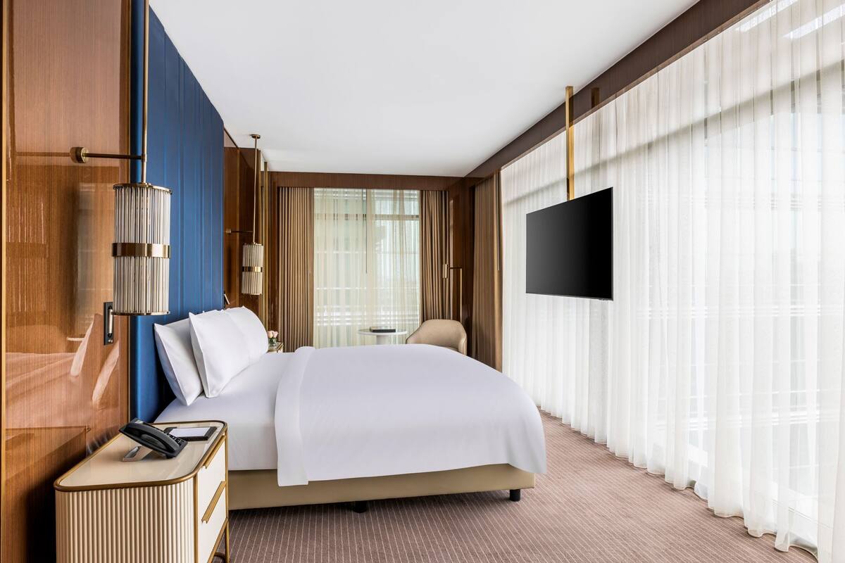A hotel room with a bed, a bedside table with a telephone, a TV mounted on the wall, and a window with sheer curtains.