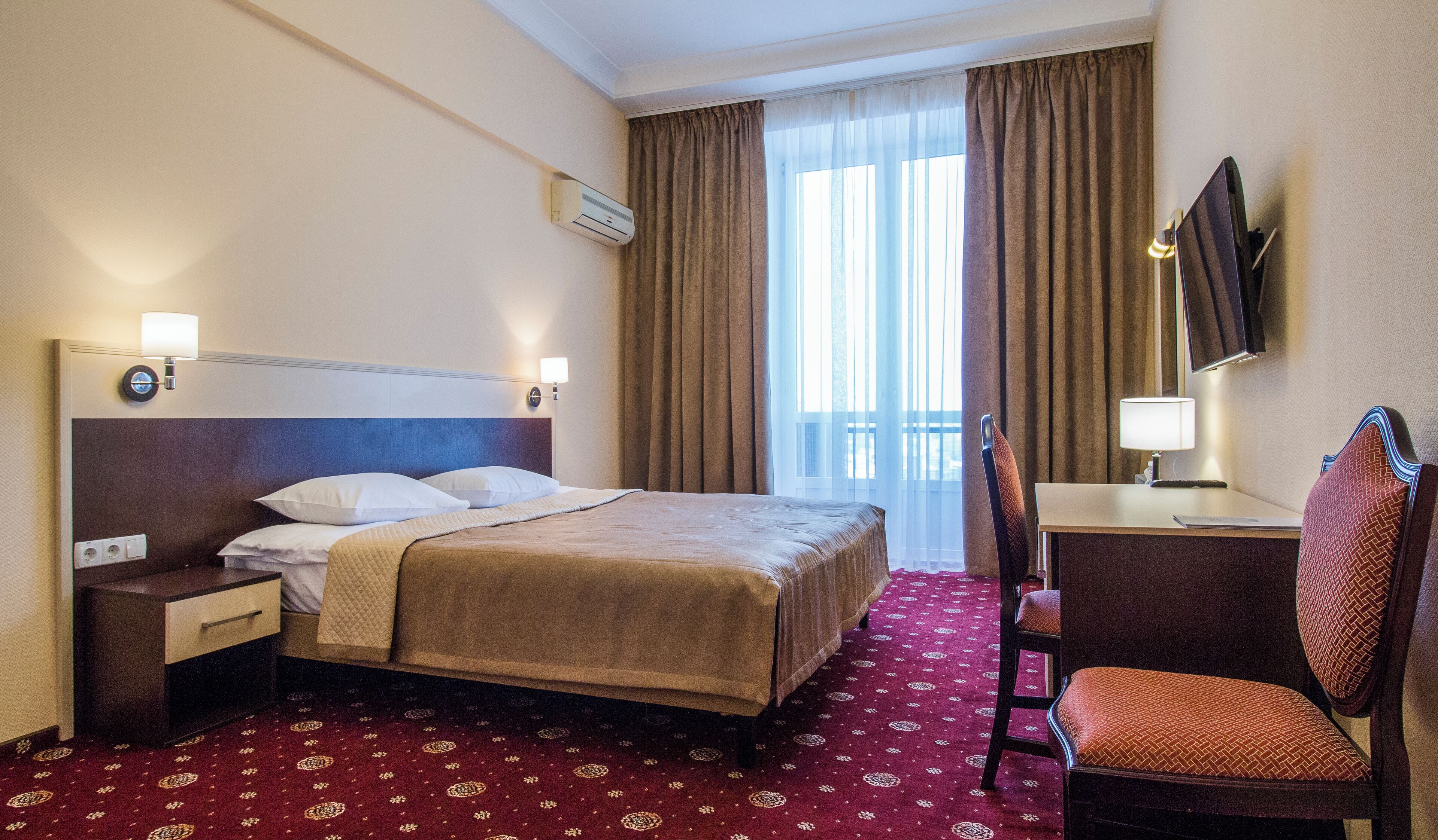 Une chambre d’hôtel équipée d’un lit, d’un bureau, d’une chaise, d’un téléviseur fixé au mur et d’une fenêtre avec des rideaux.