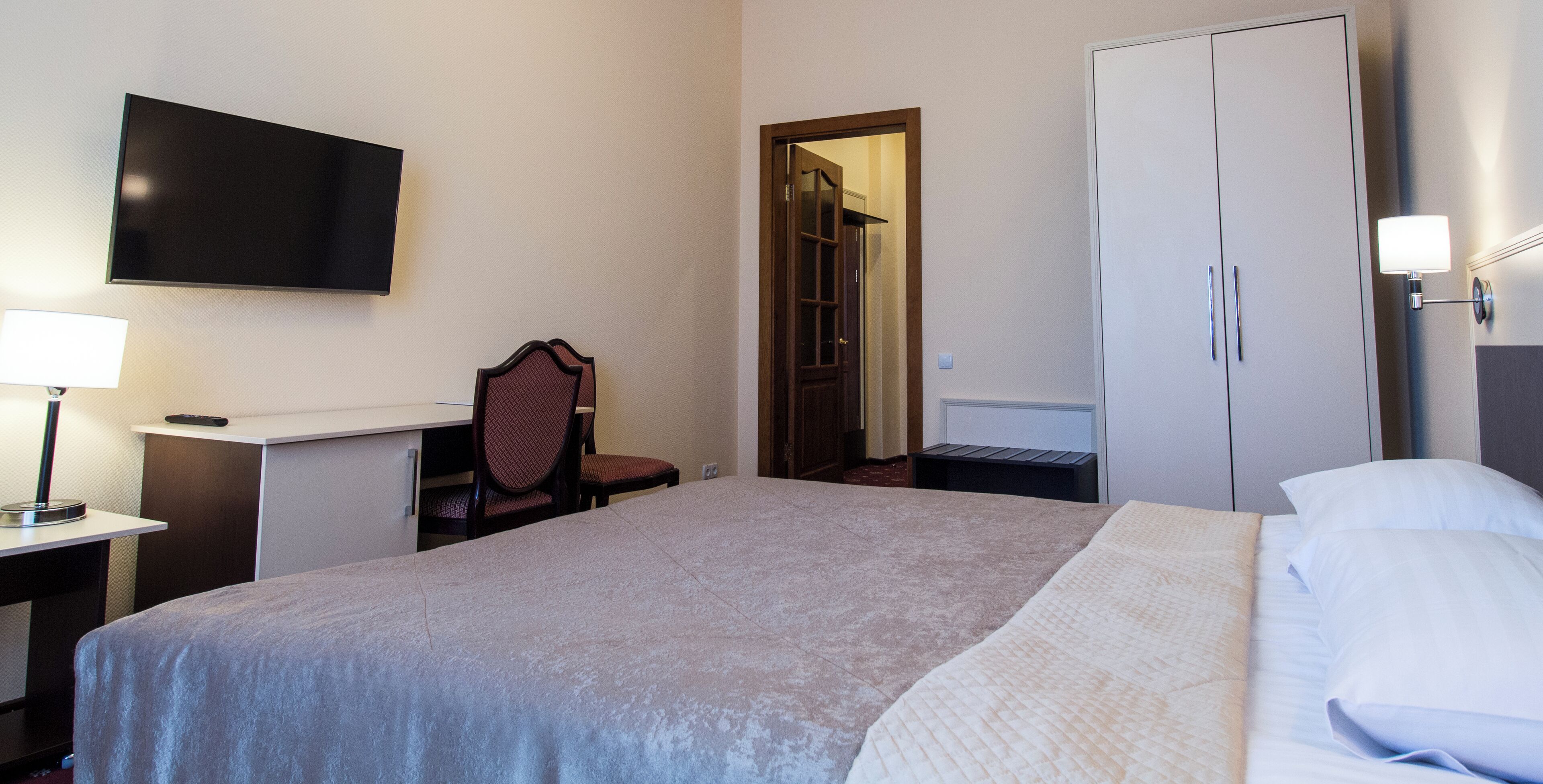 A hotel room with a bed, a desk with a lamp, a TV mounted on the wall, and a wardrobe.