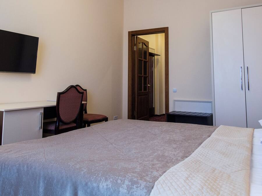 A hotel room with a bed, a desk with a lamp, a TV mounted on the wall, and a wardrobe.